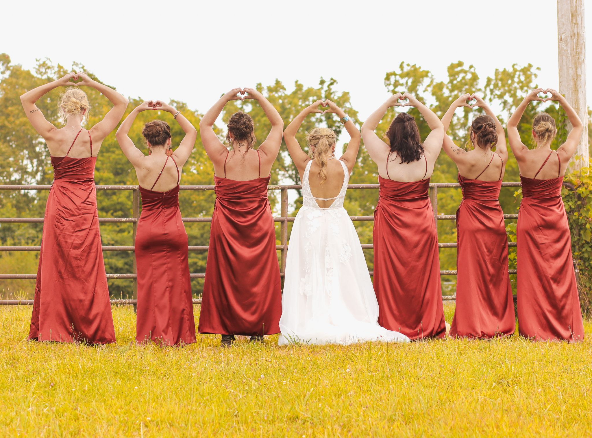 Wedding party in red dresses and white gown posing with raised bouquets against golden field.