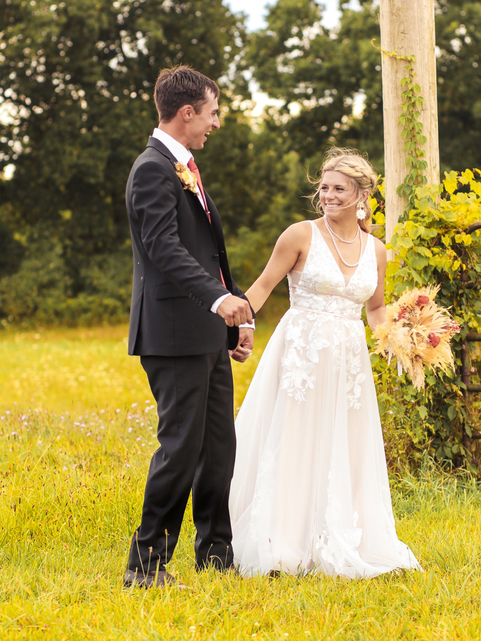 Couple in wedding attire standing together in a sunlit field with yellow flowers.