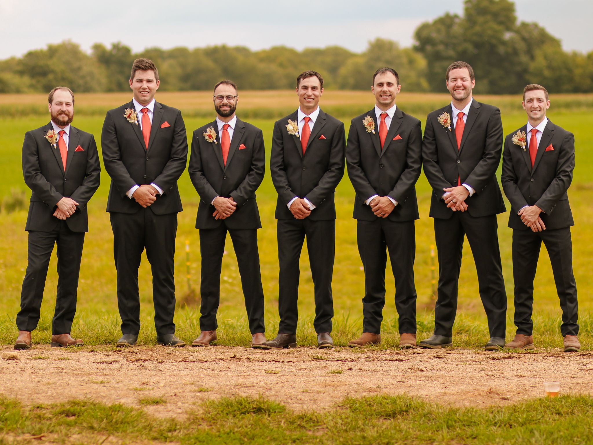 Groomsmen in dark suits with red ties stand in line outdoors against natural landscape.