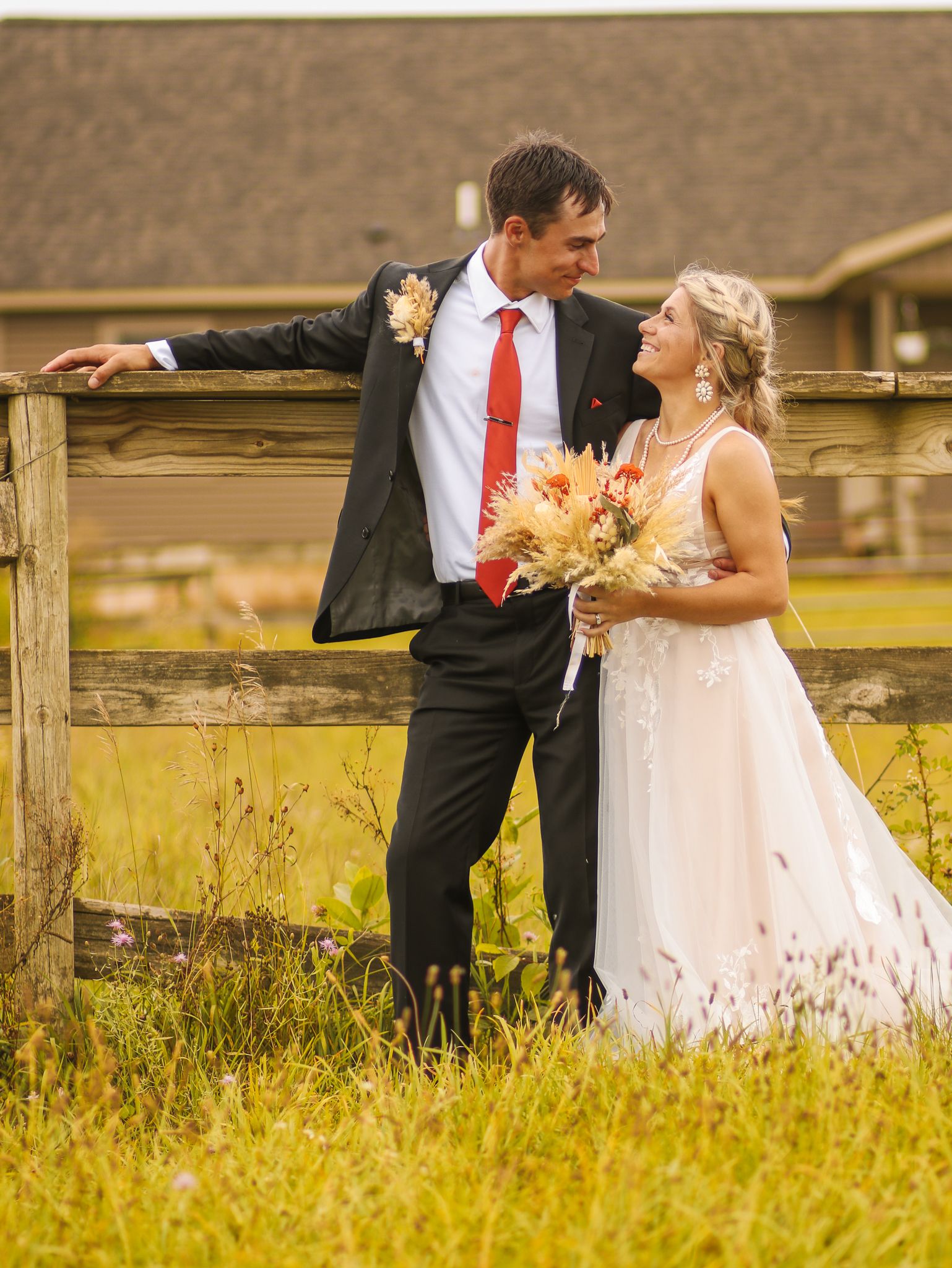 Newlyweds pose by wooden fence in rustic outdoor setting with golden grass.
