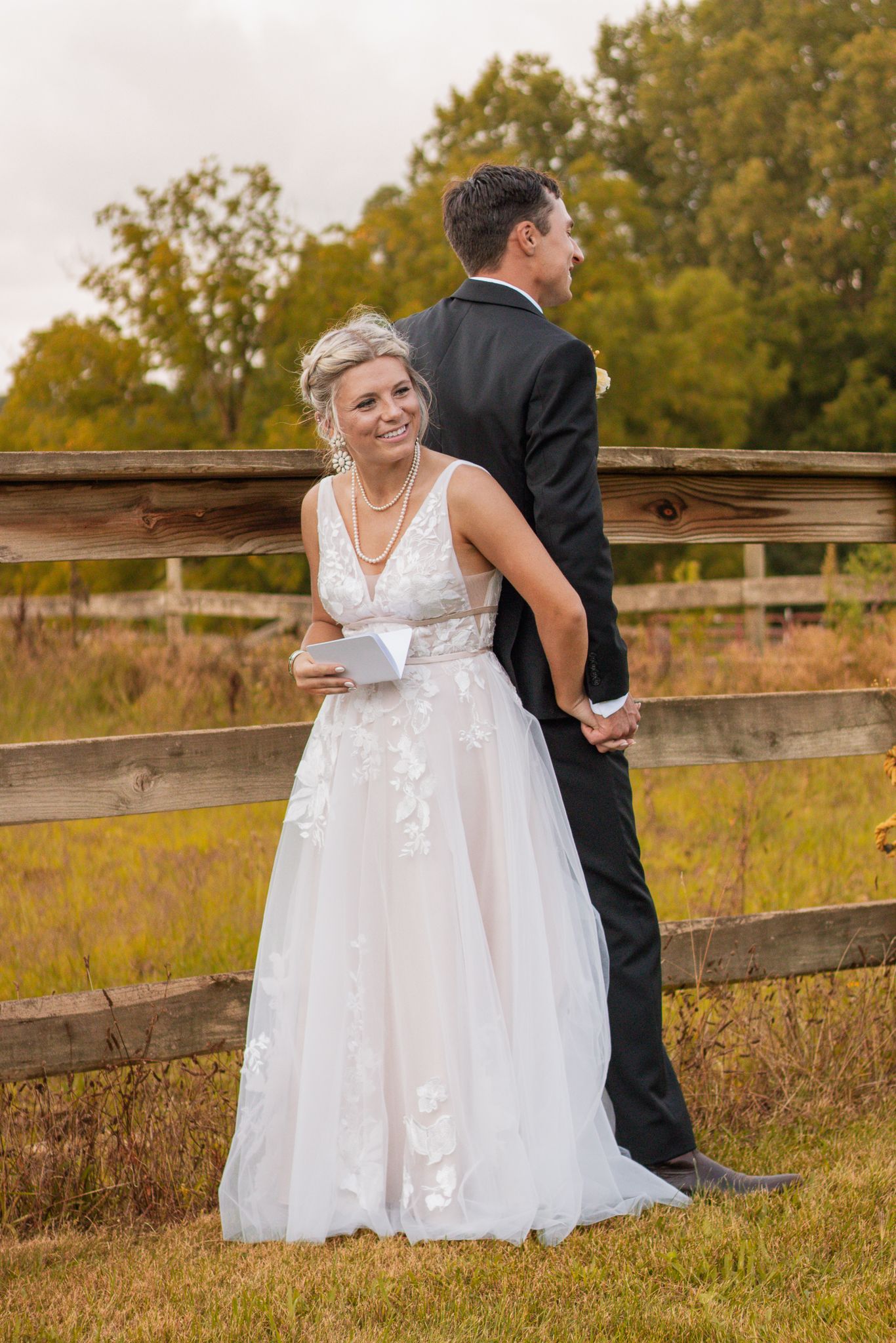 Wedding couple poses against wooden fence with autumn field landscape in background.