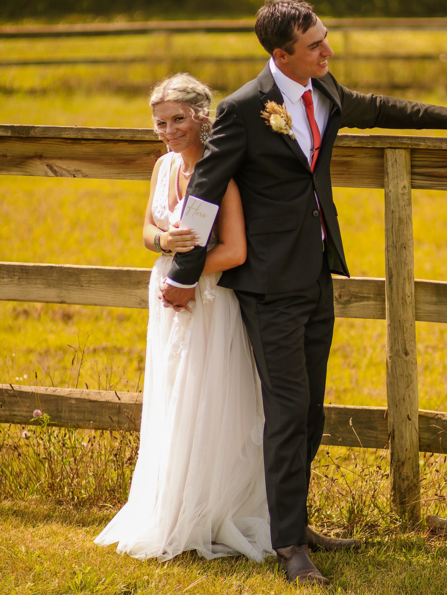 Newlyweds embrace while standing against rustic fence with golden field in background.
