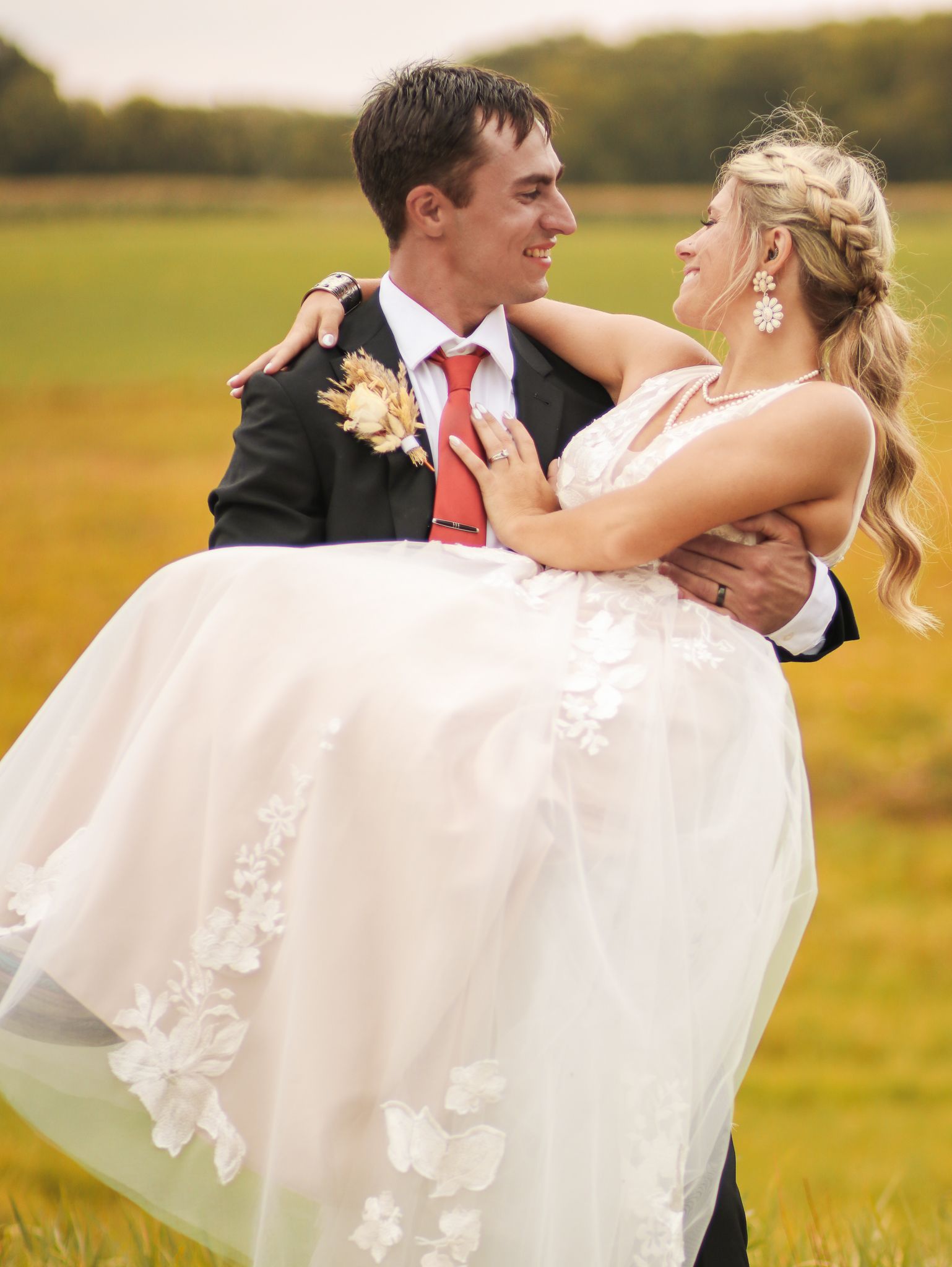 Groom carries bride in white dress against golden field background.