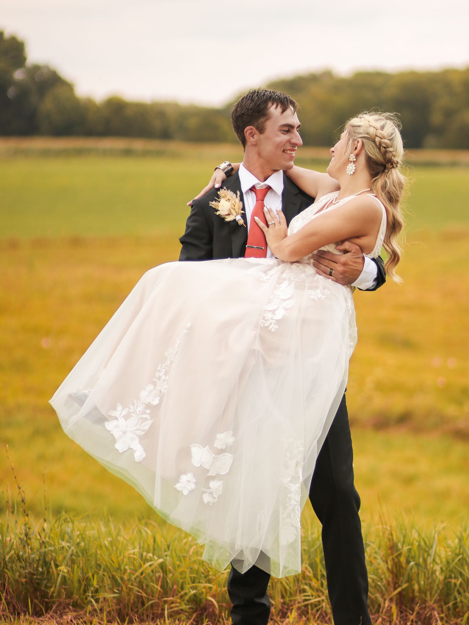 Happy couple sharing romantic moment with groom carrying bride in field setting.
