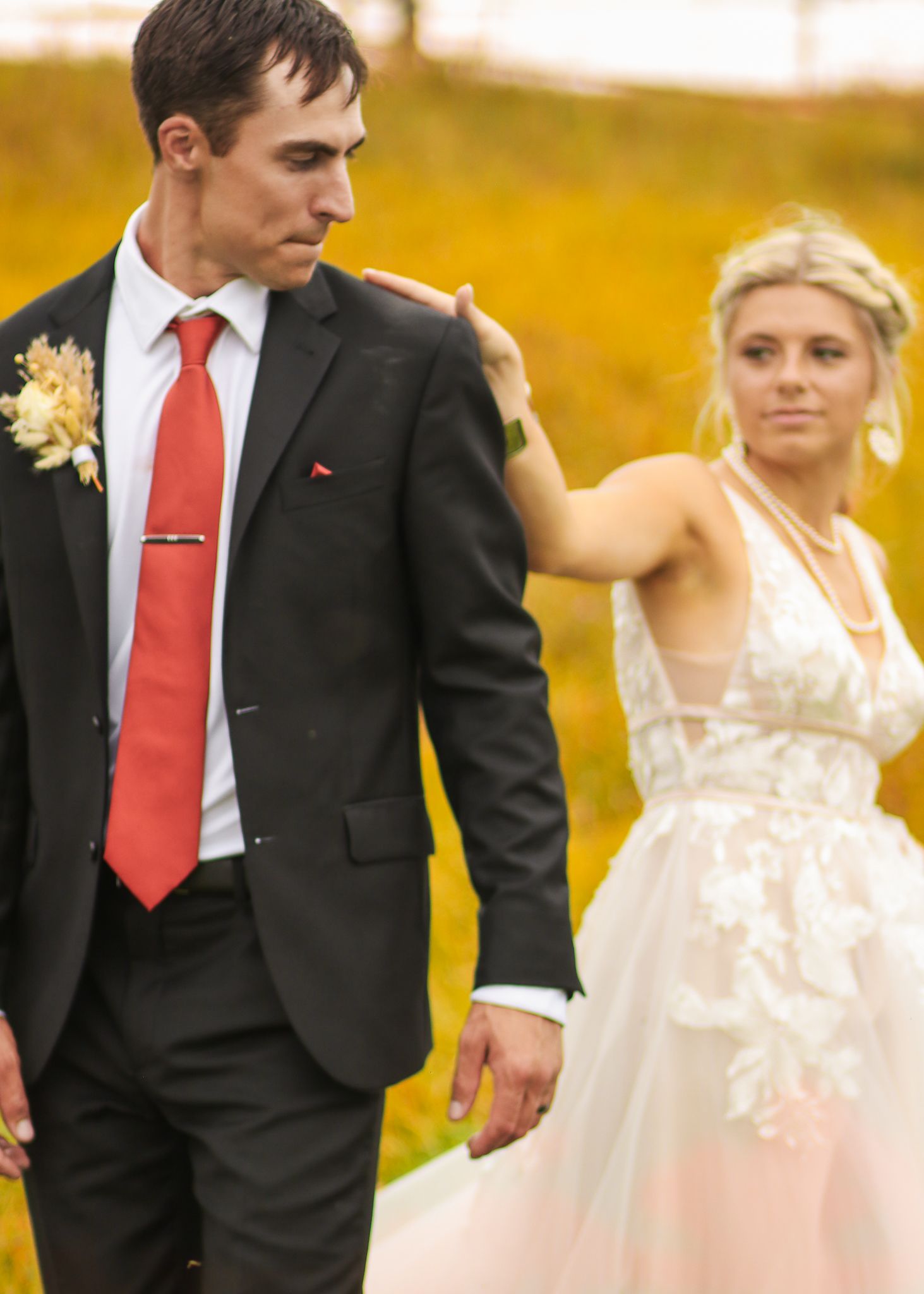 Newlyweds pose together in a golden field during sunset, with the bride in white and groom in dark suit with red tie.