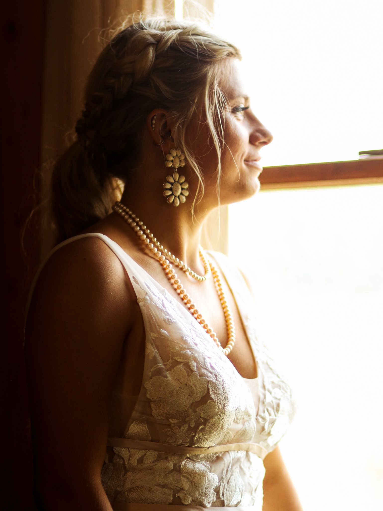 Bridal portrait in silhouette showing lace dress details and pearl accessories near window.
