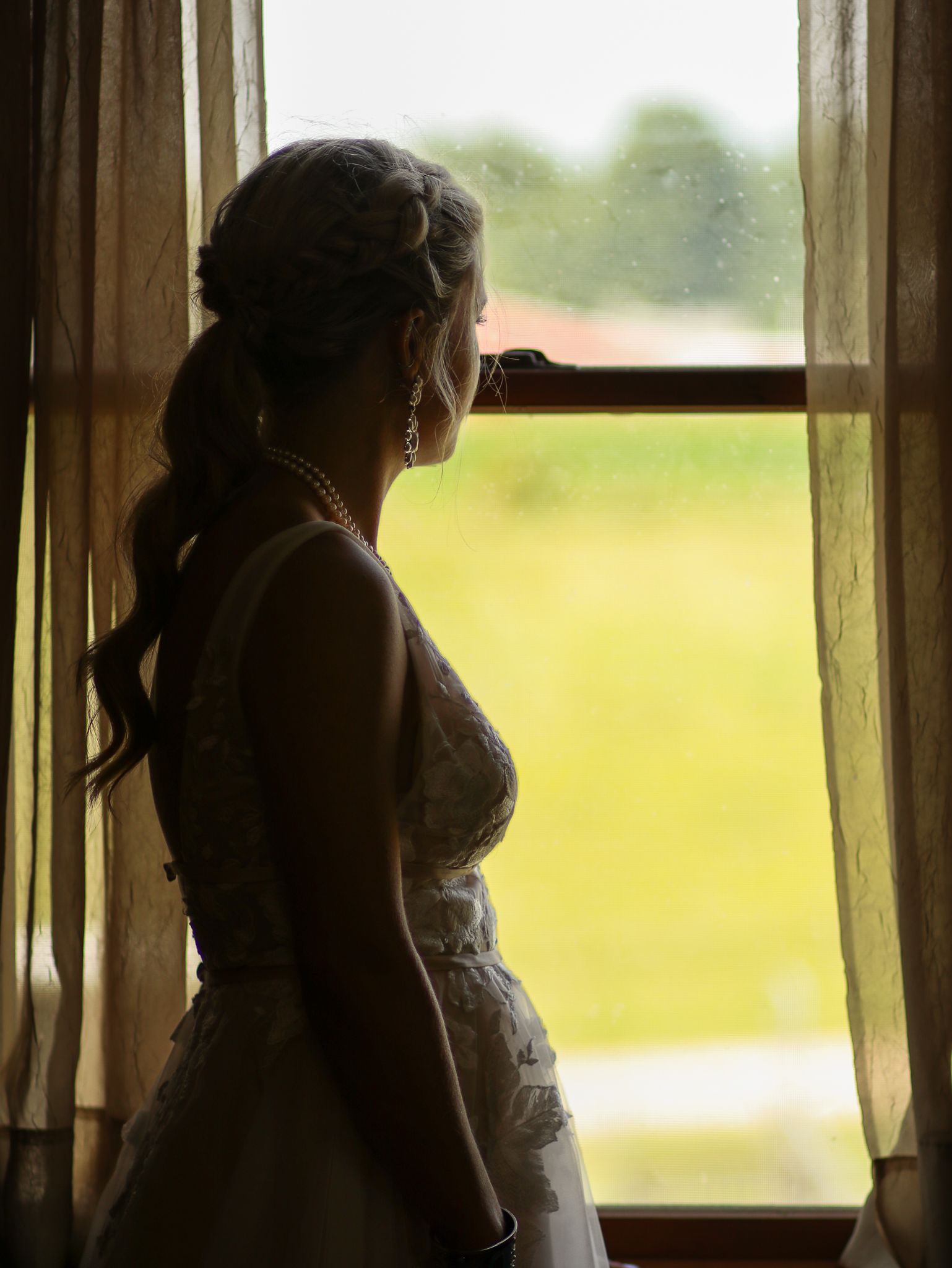 A silhouetted figure in a wedding dress stands beside a window with sheer curtains and sunlit scenery outside.
