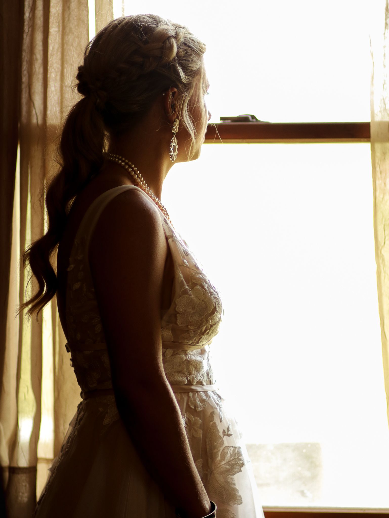 Silhouette of a bride in a lace dress wearing pearl earrings stands by a sunlit window.