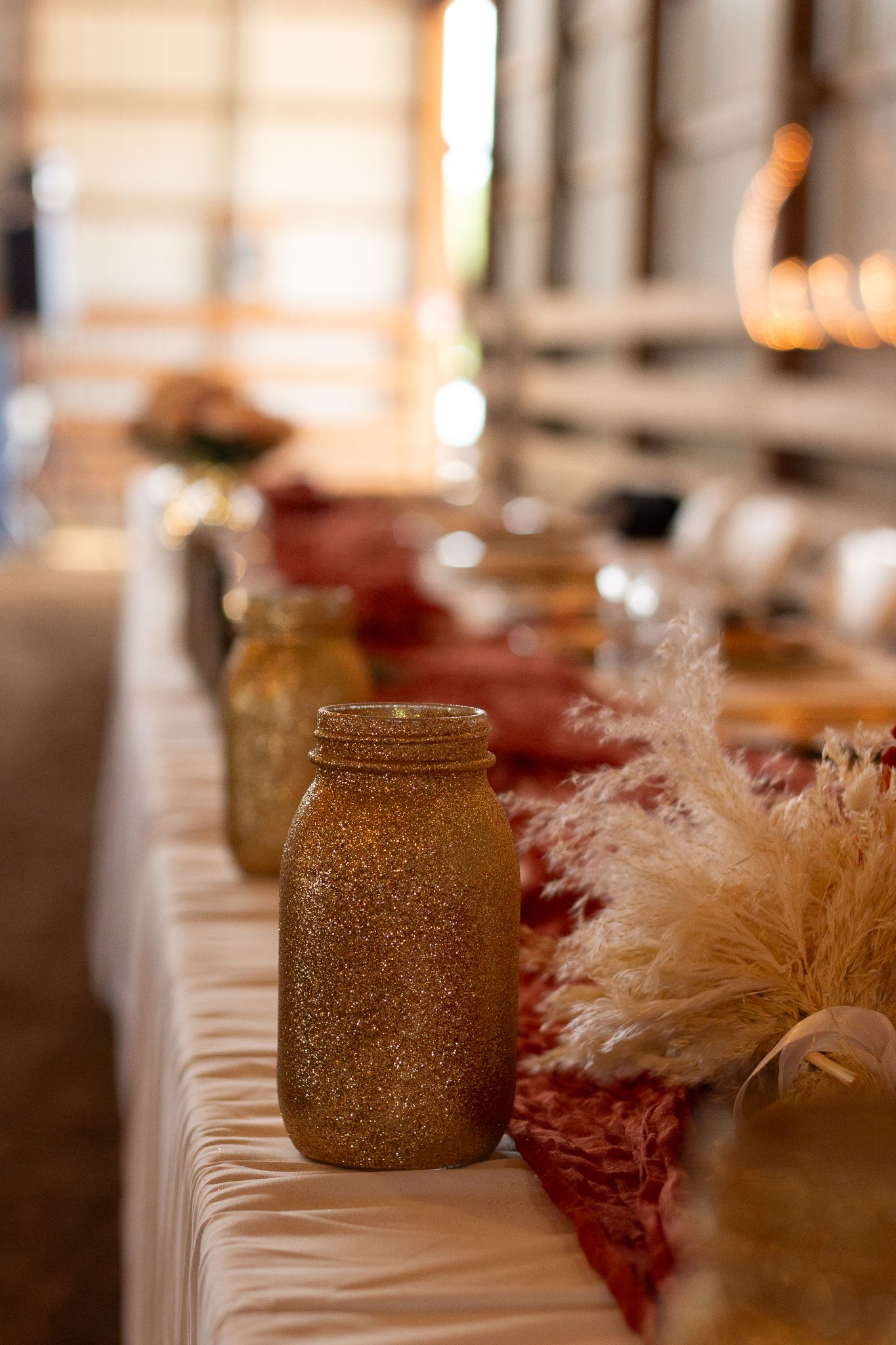 Glittery mason jars and pampas grass decorate a white-clothed table at a rustic wedding reception.