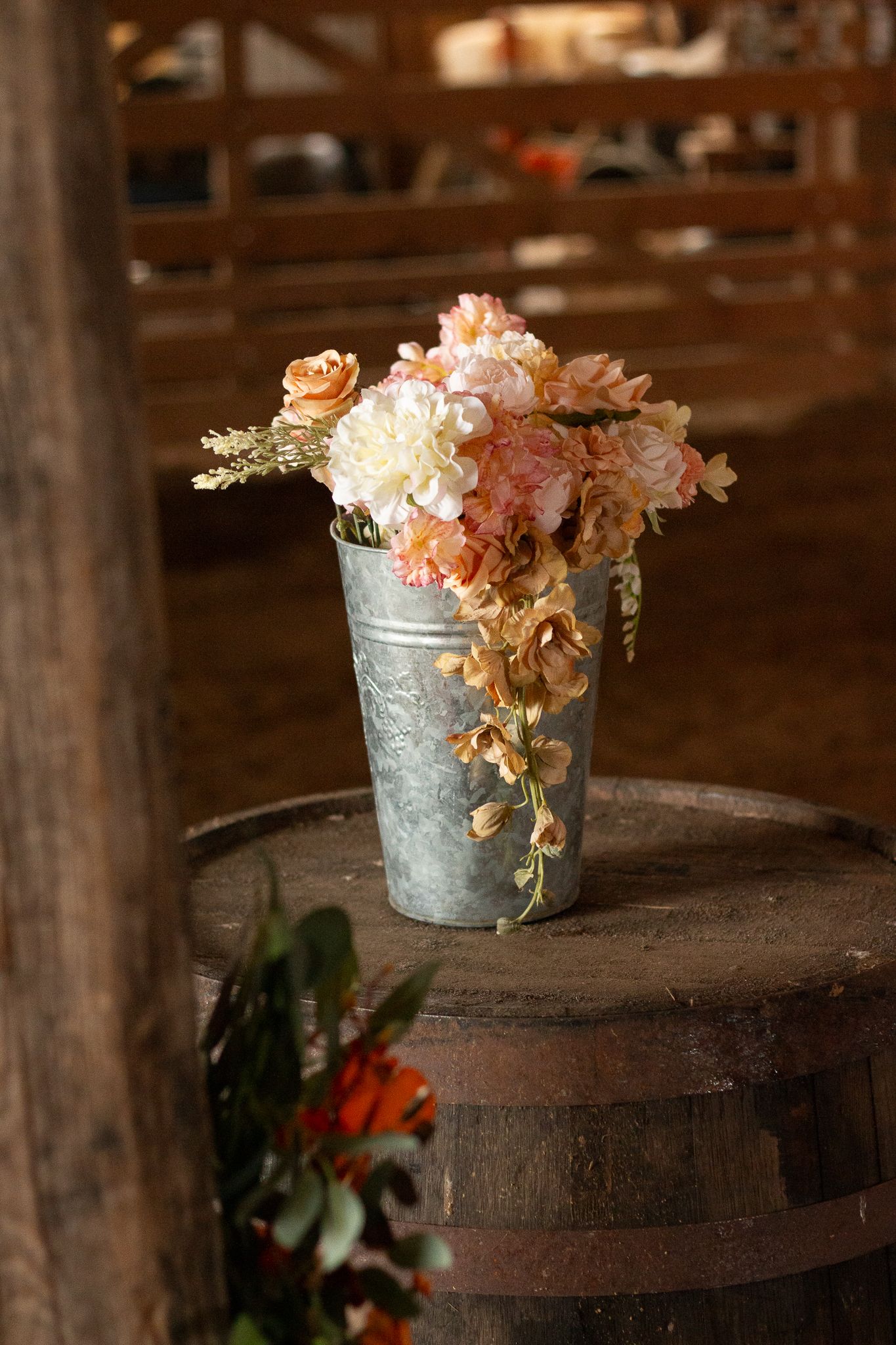Rustic floral arrangement of pink and white flowers in a galvanized metal bucket on wooden barrel.