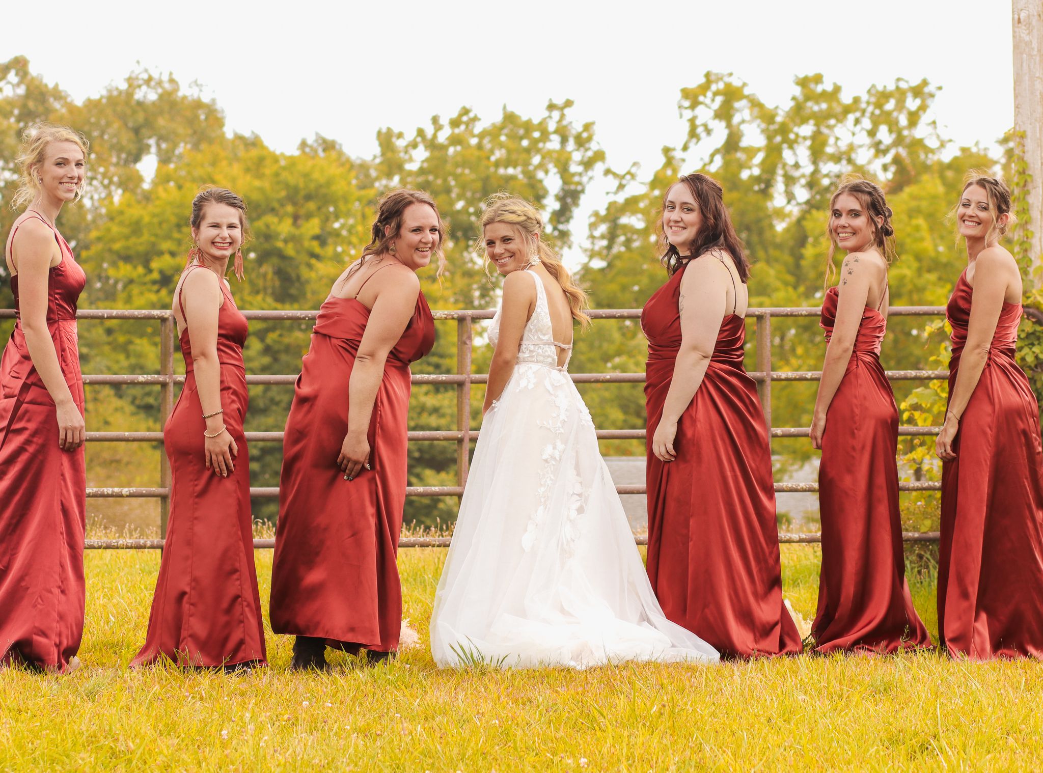 Bridesmaids in red dresses pose with bride in white gown against wooden fence backdrop.