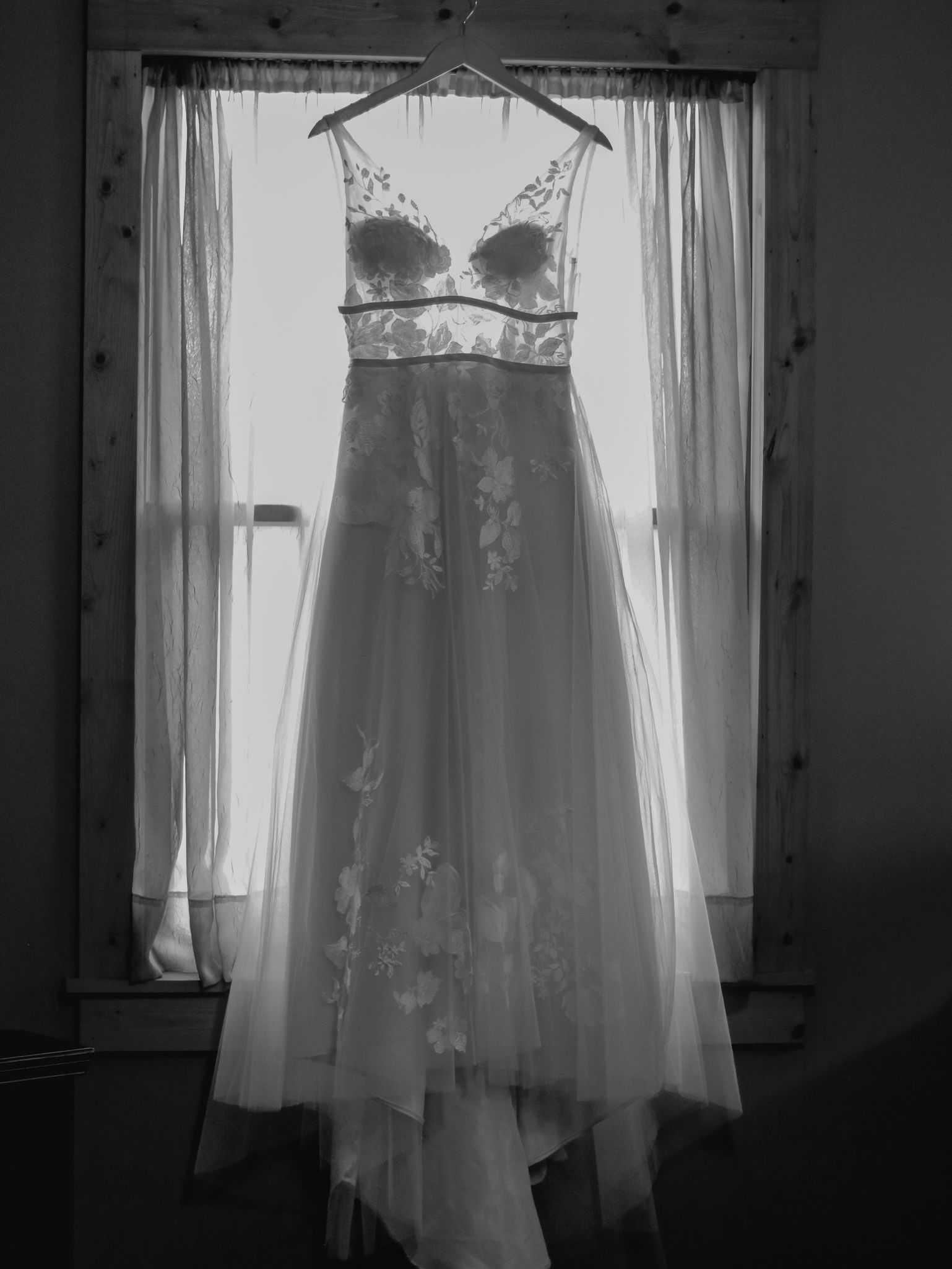 White lace wedding dress hanging in window light creating a romantic silhouette in black and white.
