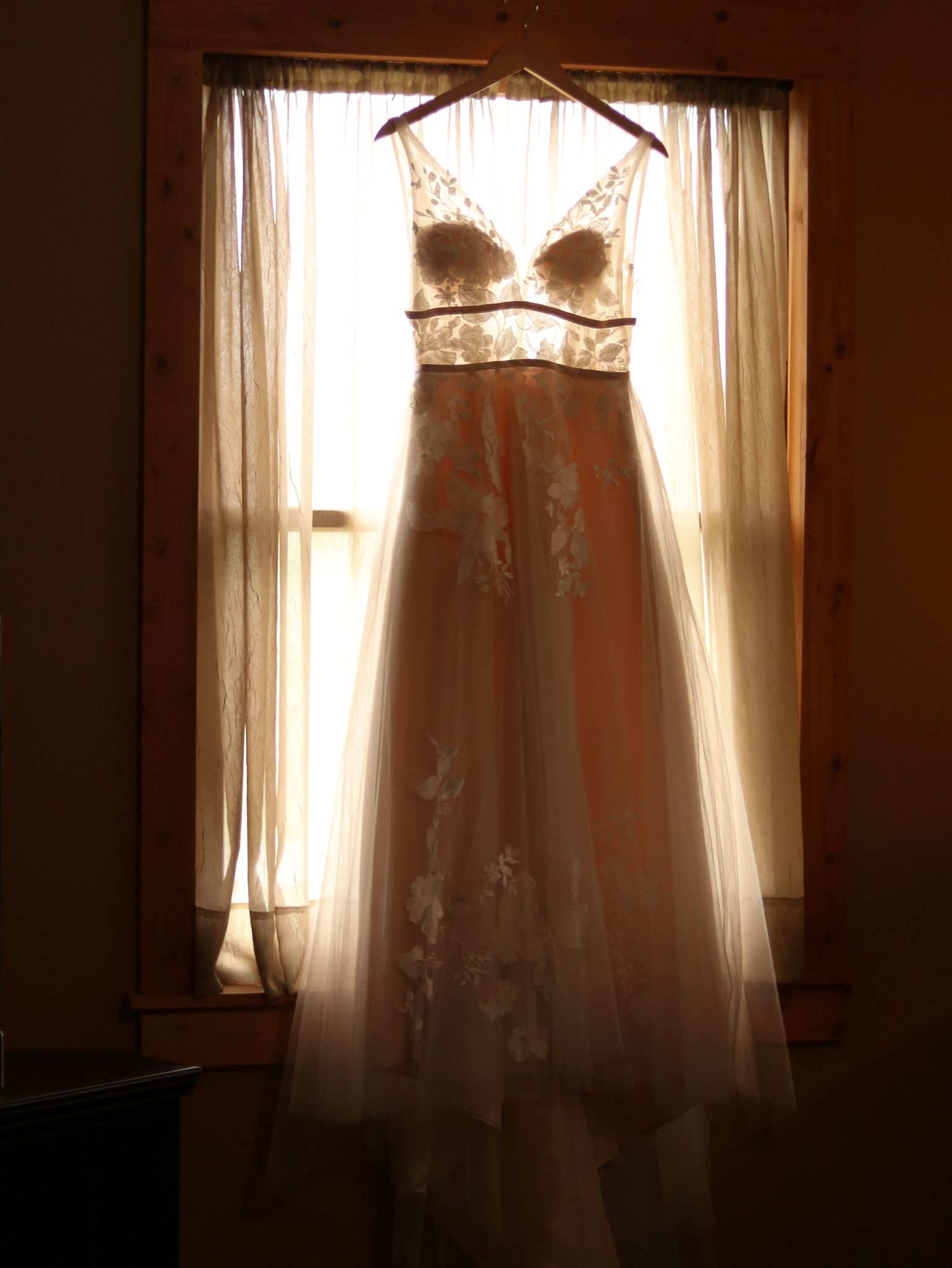 Ethereal wedding dress with lace details backlit by sunlight through window curtains.