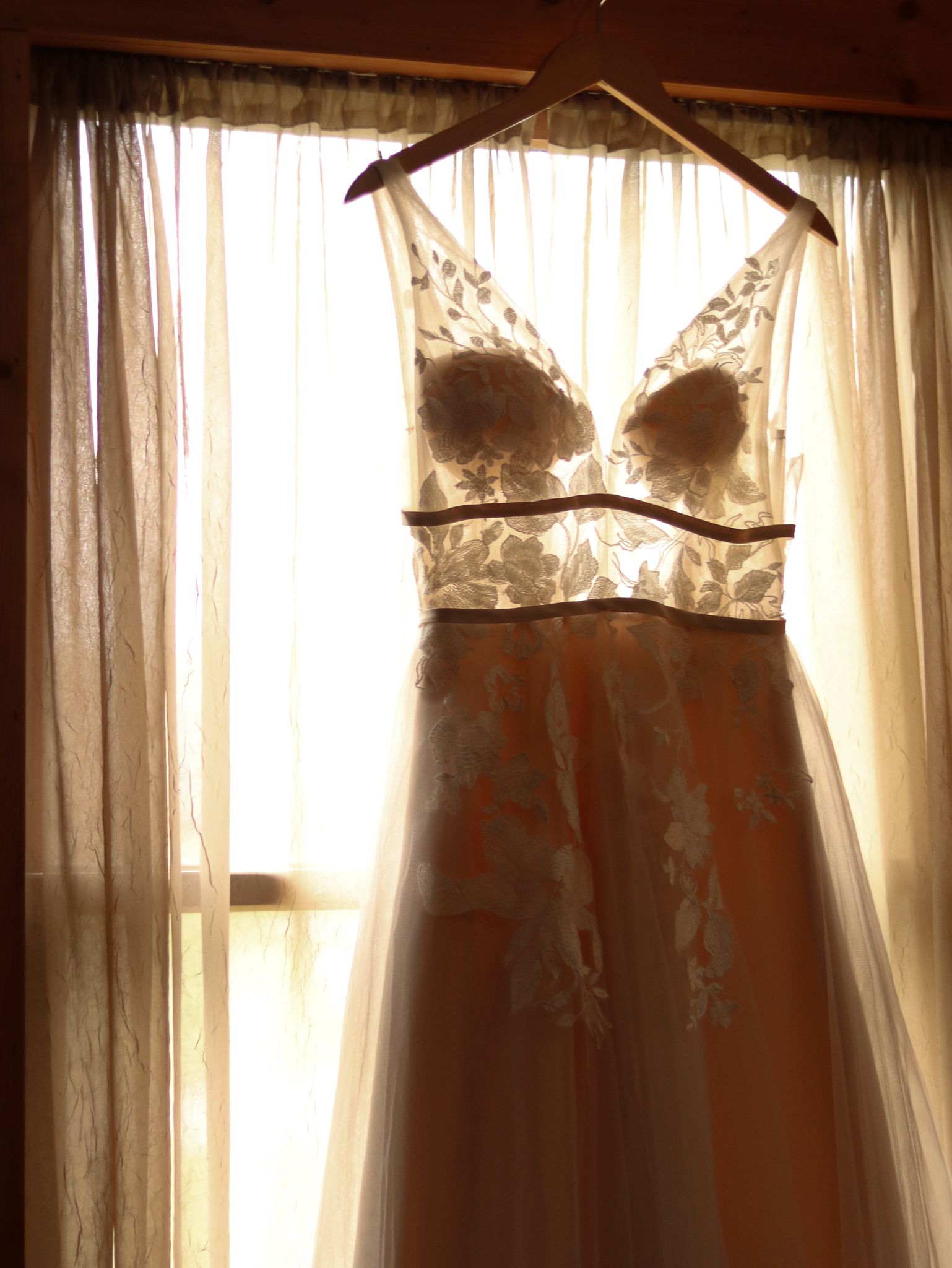 An elegant white wedding dress with lace bodice hangs by a window with sheer curtains in golden sunlight.