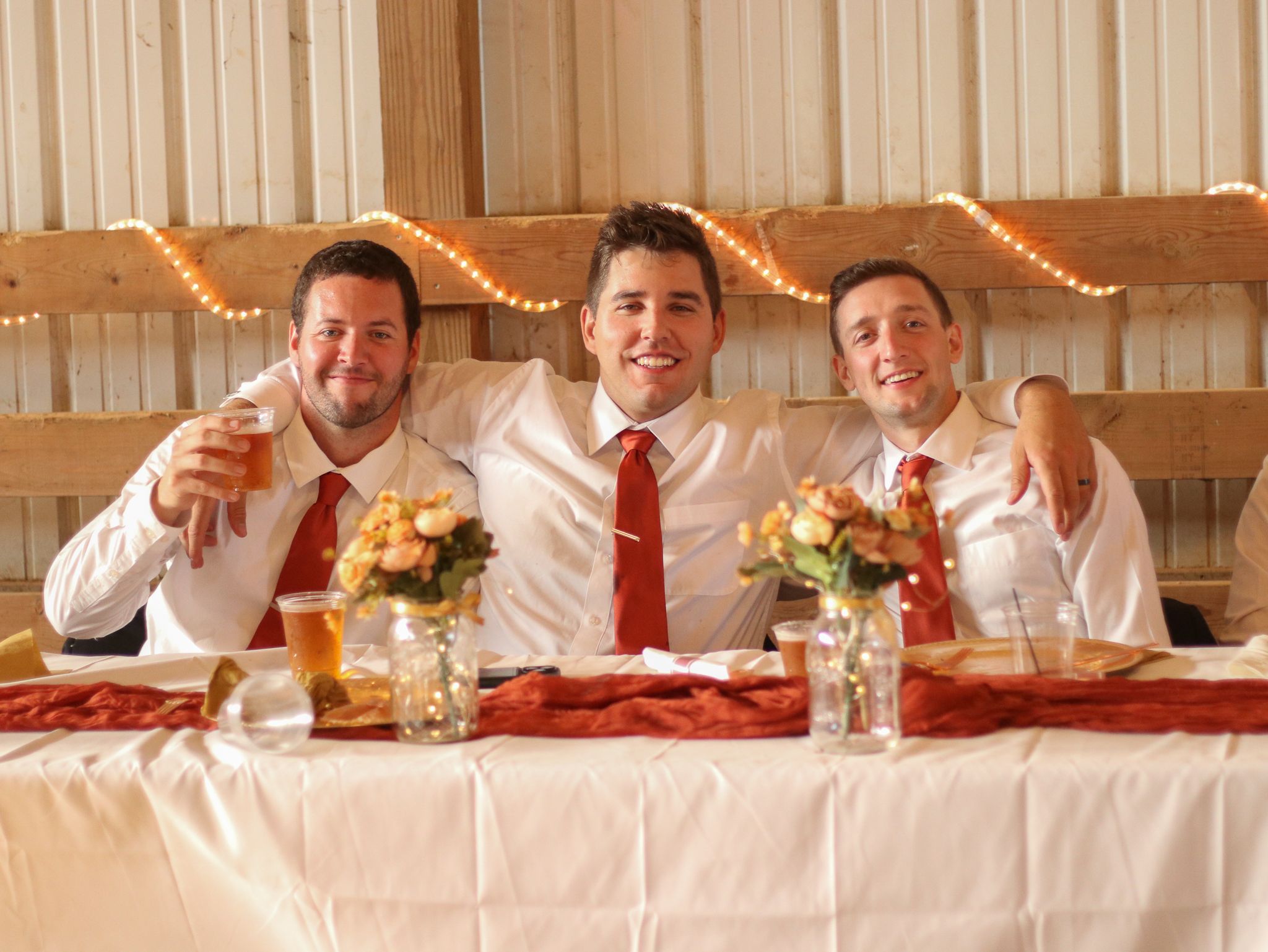 Three groomsmen in white shirts and red ties toast at wedding reception head table.