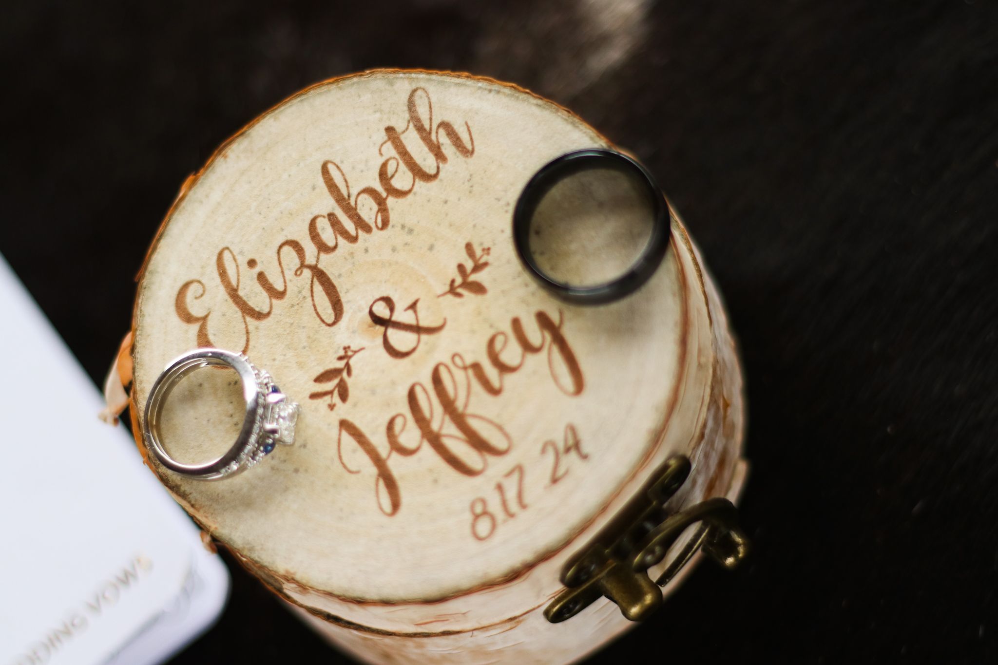 Wedding rings displayed on a rustic wooden ring box engraved with names and a date.