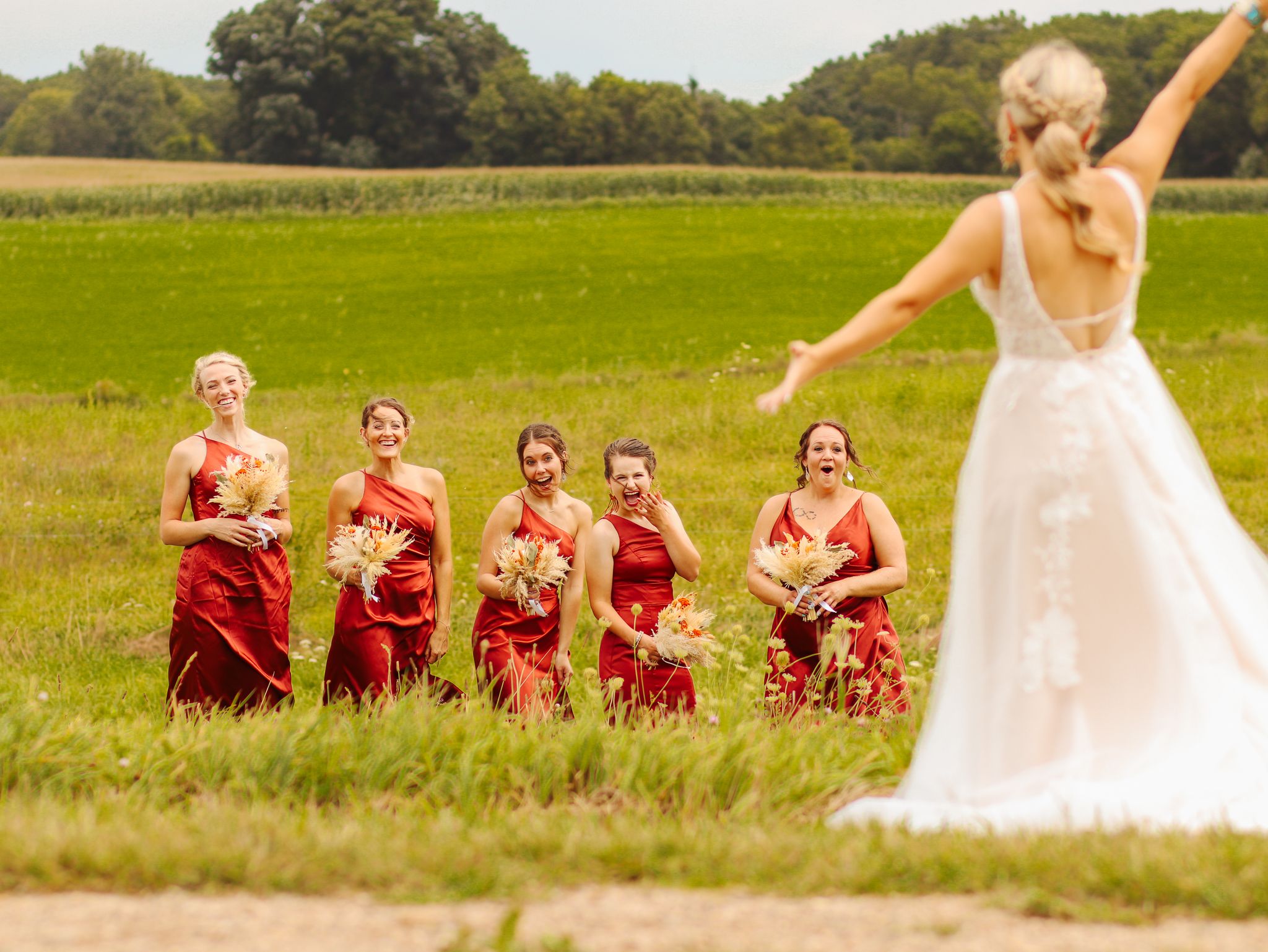 Bridesmaids in red dresses stand together in a field as bride poses in white wedding gown.