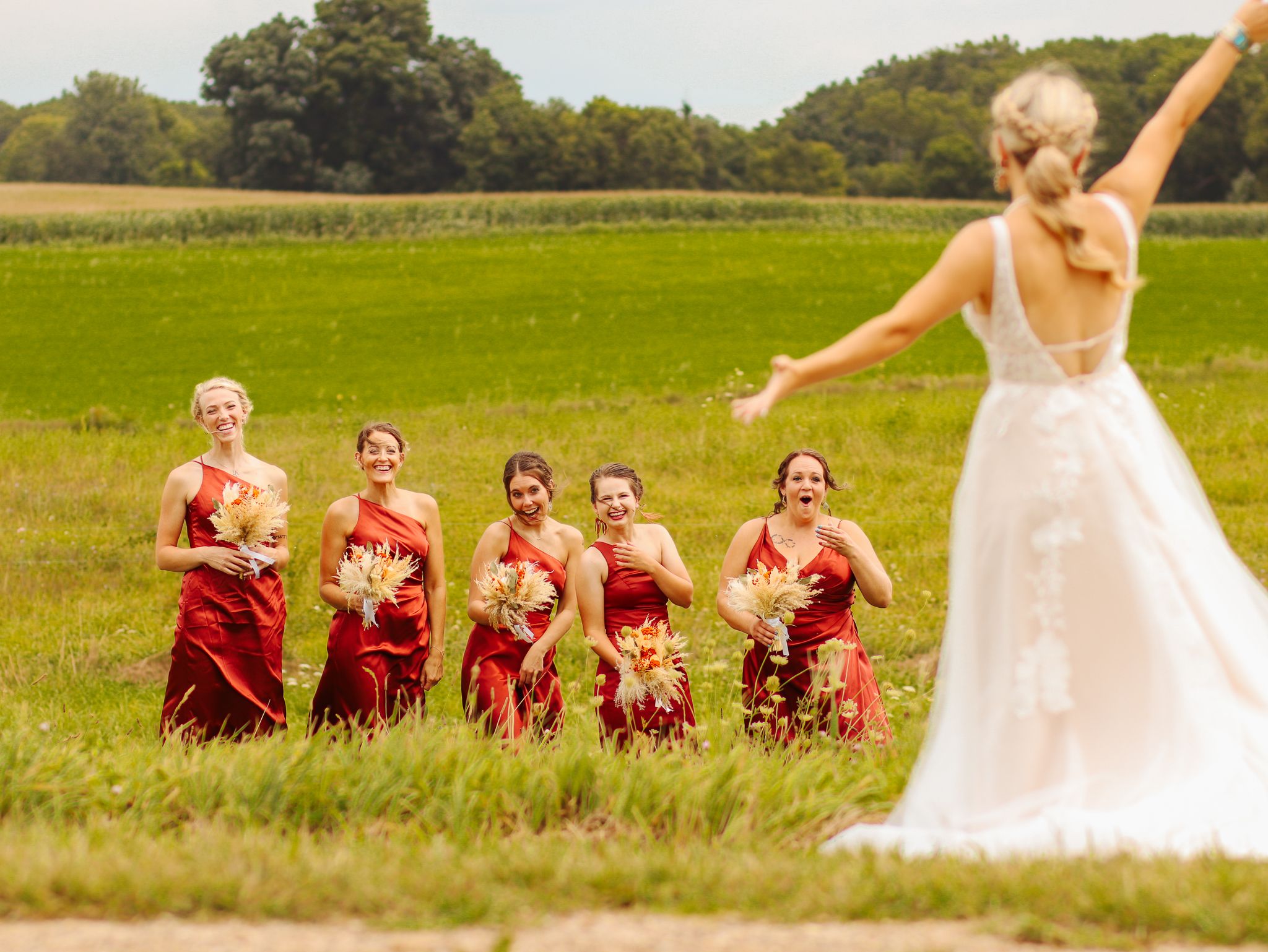 Candid wedding photo showing bridesmaids in red dresses reacting to bride's playful pose in field.