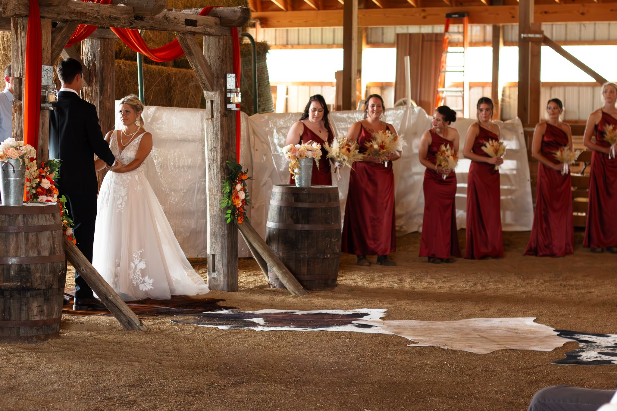 A rustic barn wedding ceremony with burgundy-dressed attendants watching in the background.