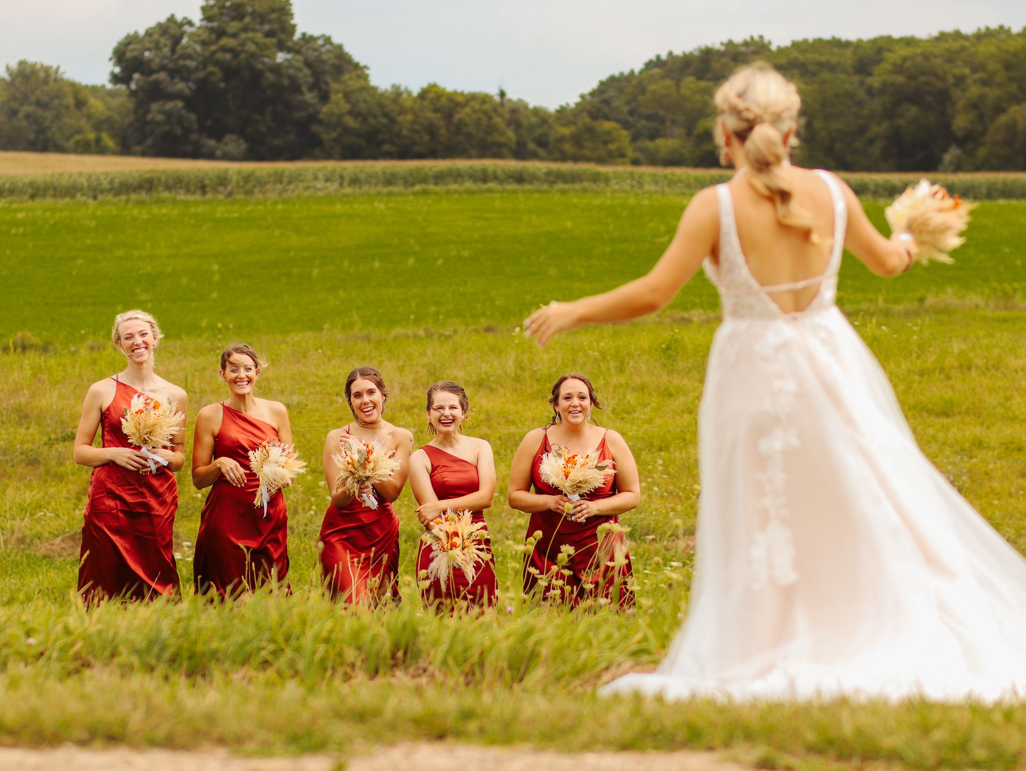 A bride in white spins joyfully in a field while bridesmaids in red dresses look on.