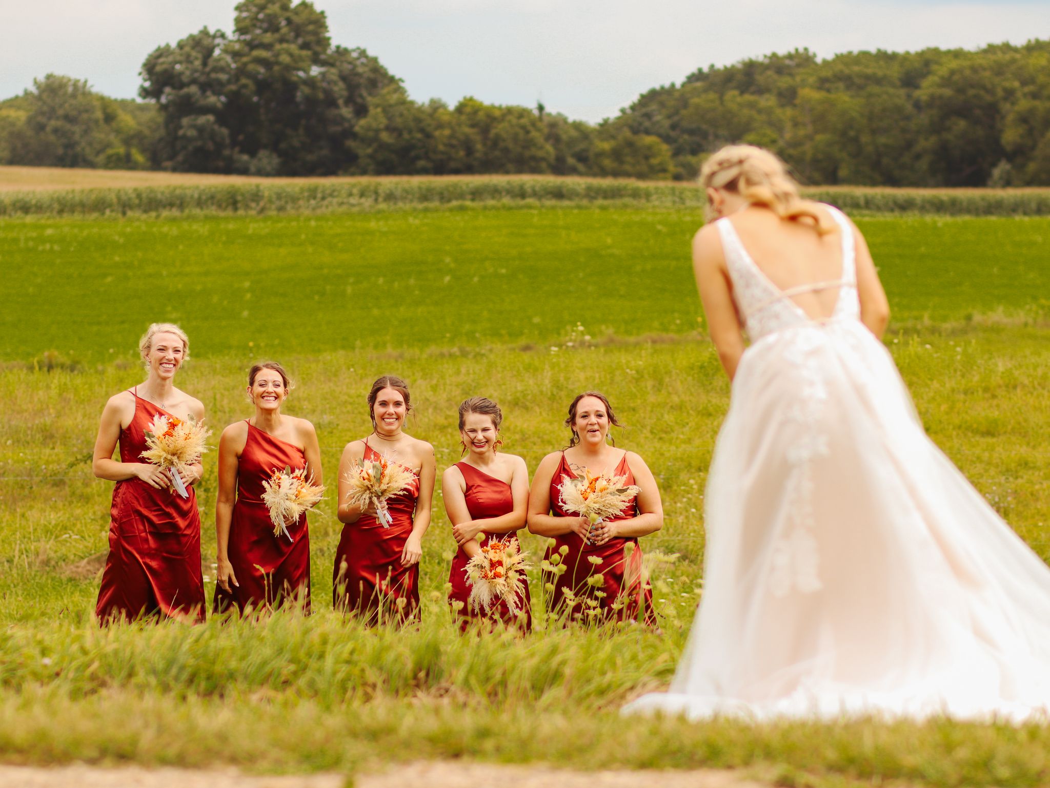 A sunny outdoor wedding scene with bridesmaids in red watching the bride in a grassy field.