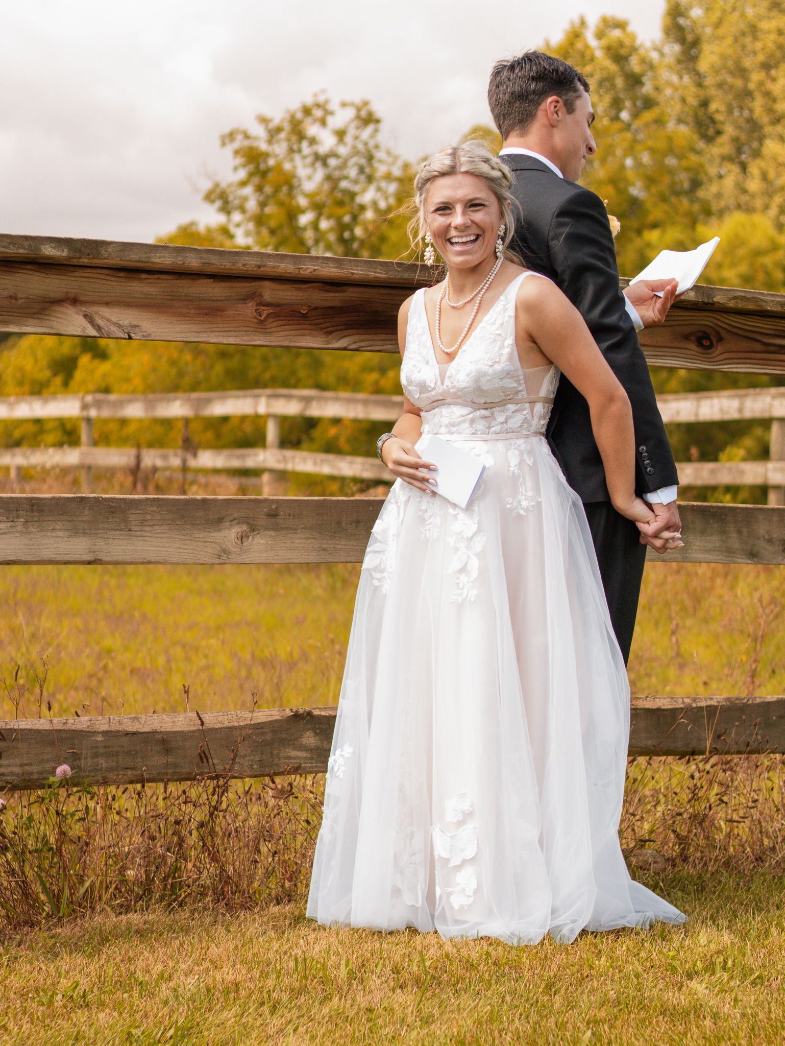 Bride in lace white gown and groom in black suit stand back-to-back at wooden fence.
