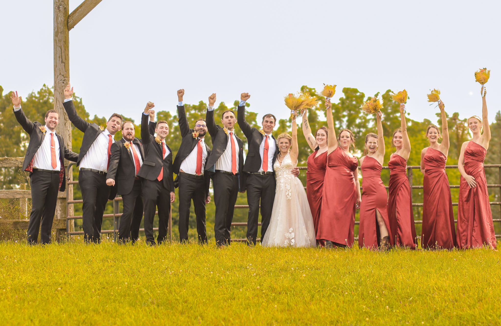 Wedding party in formal attire poses together in a yellow field with arms raised joyfully.