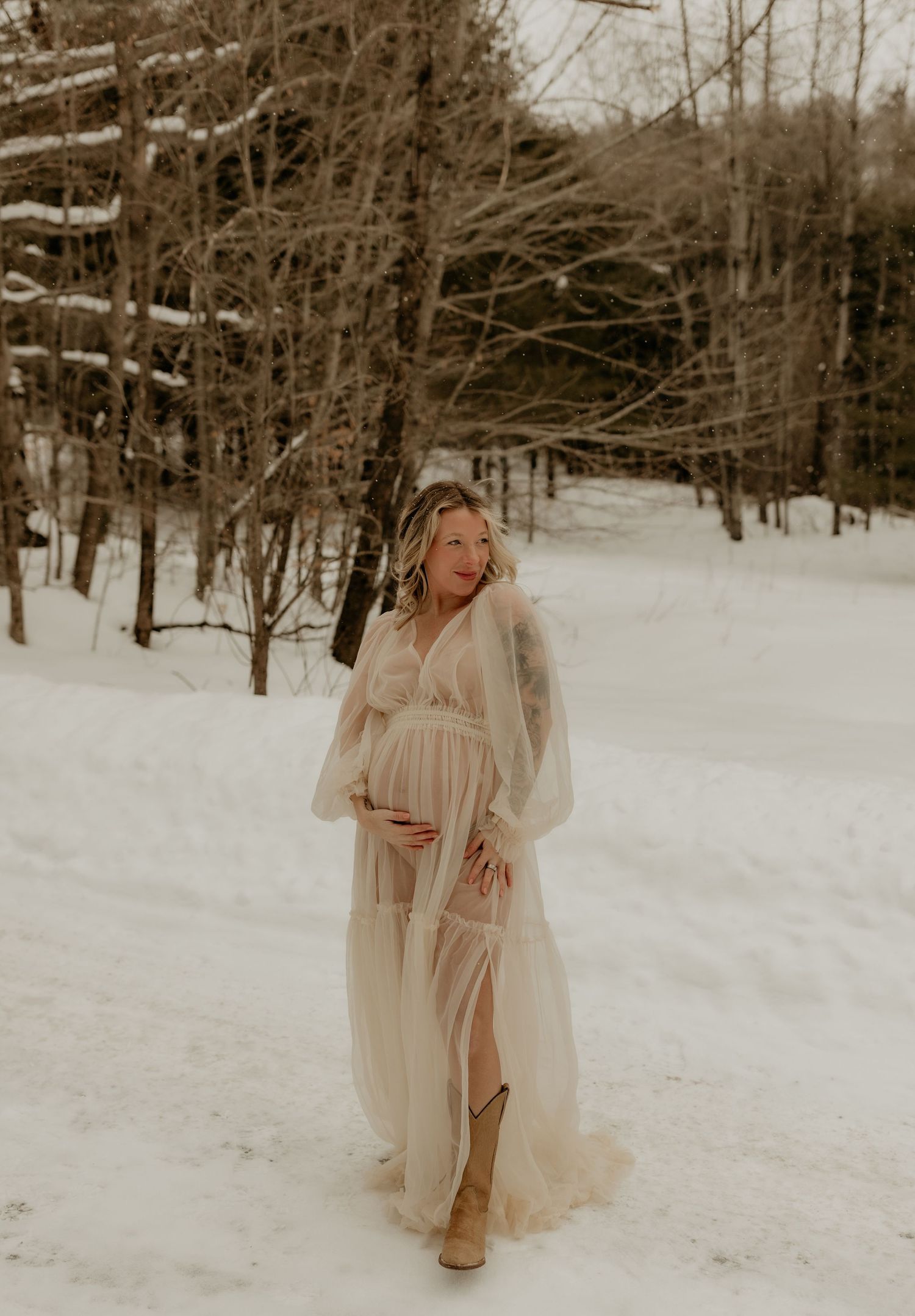 Woman in flowing cream dress poses in snowy winter forest setting.