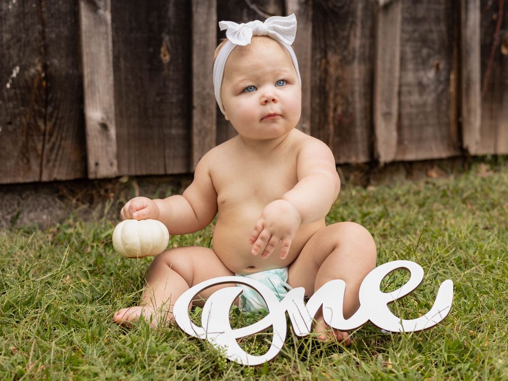 Baby wearing white bow sits on grass near wooden fence in series of candid outdoor portraits.