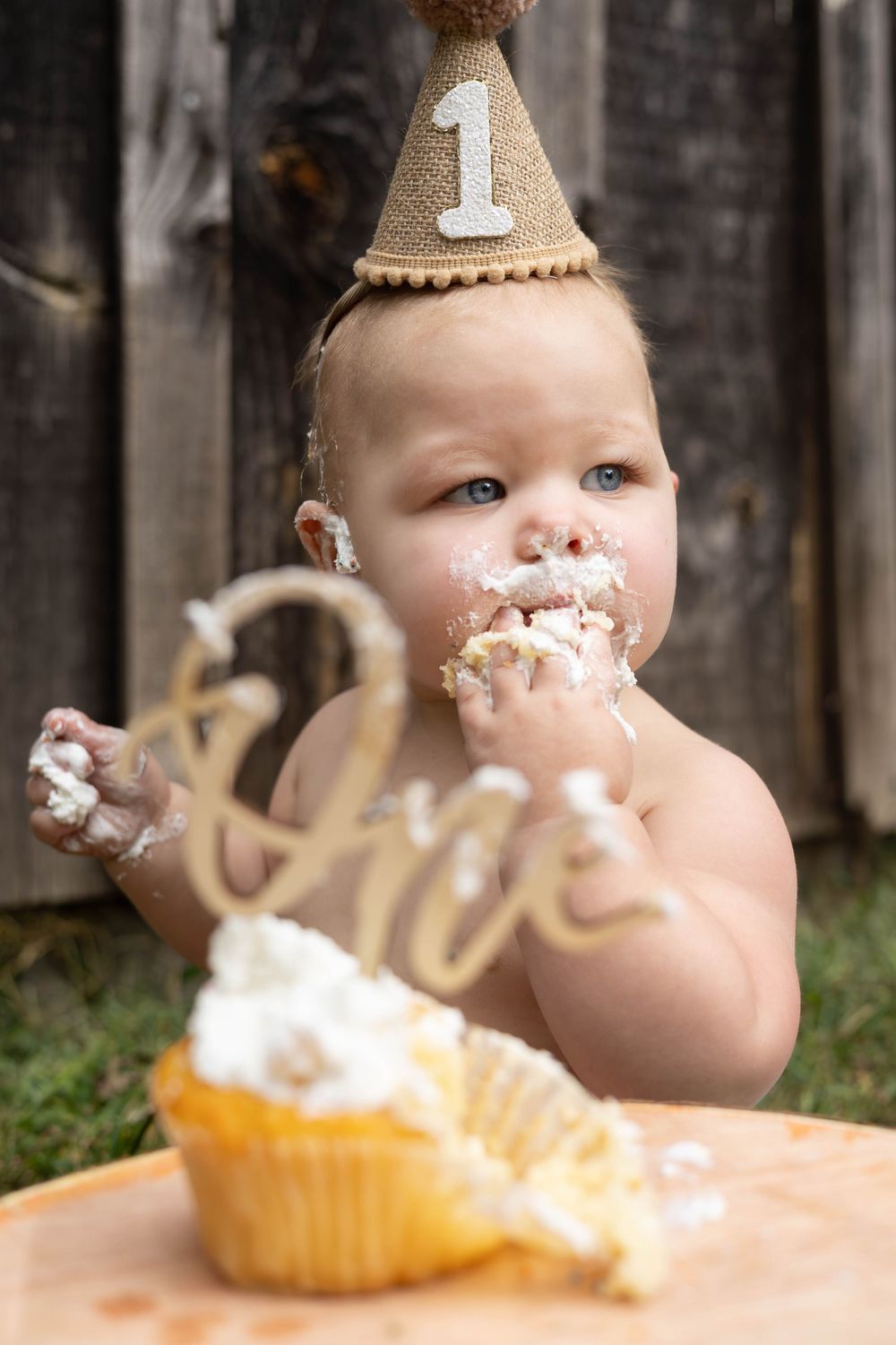 A baby in a party hat enjoys their first birthday cake with frosting smeared on their face during a cake smash photoshoot.