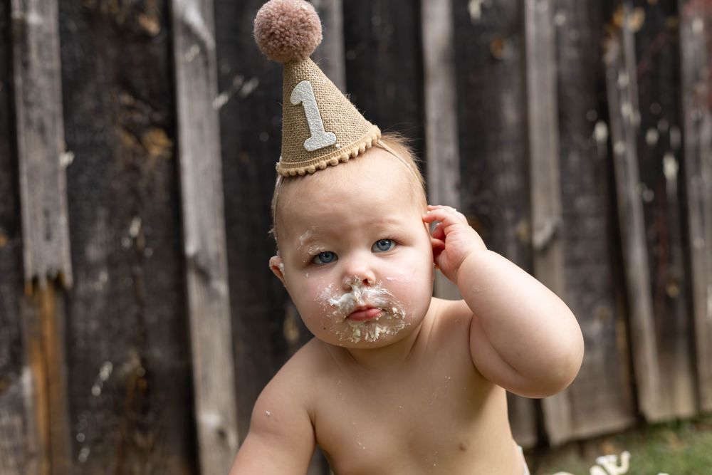 A baby wearing a pearl necklace and diaper sits outdoors near a wooden fence.
