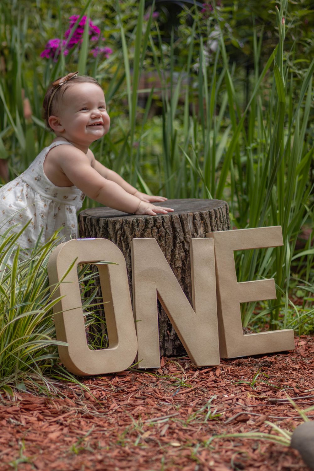 A smiling child in a white dress poses with wooden letters spelling 'ONE' on a tree stump.