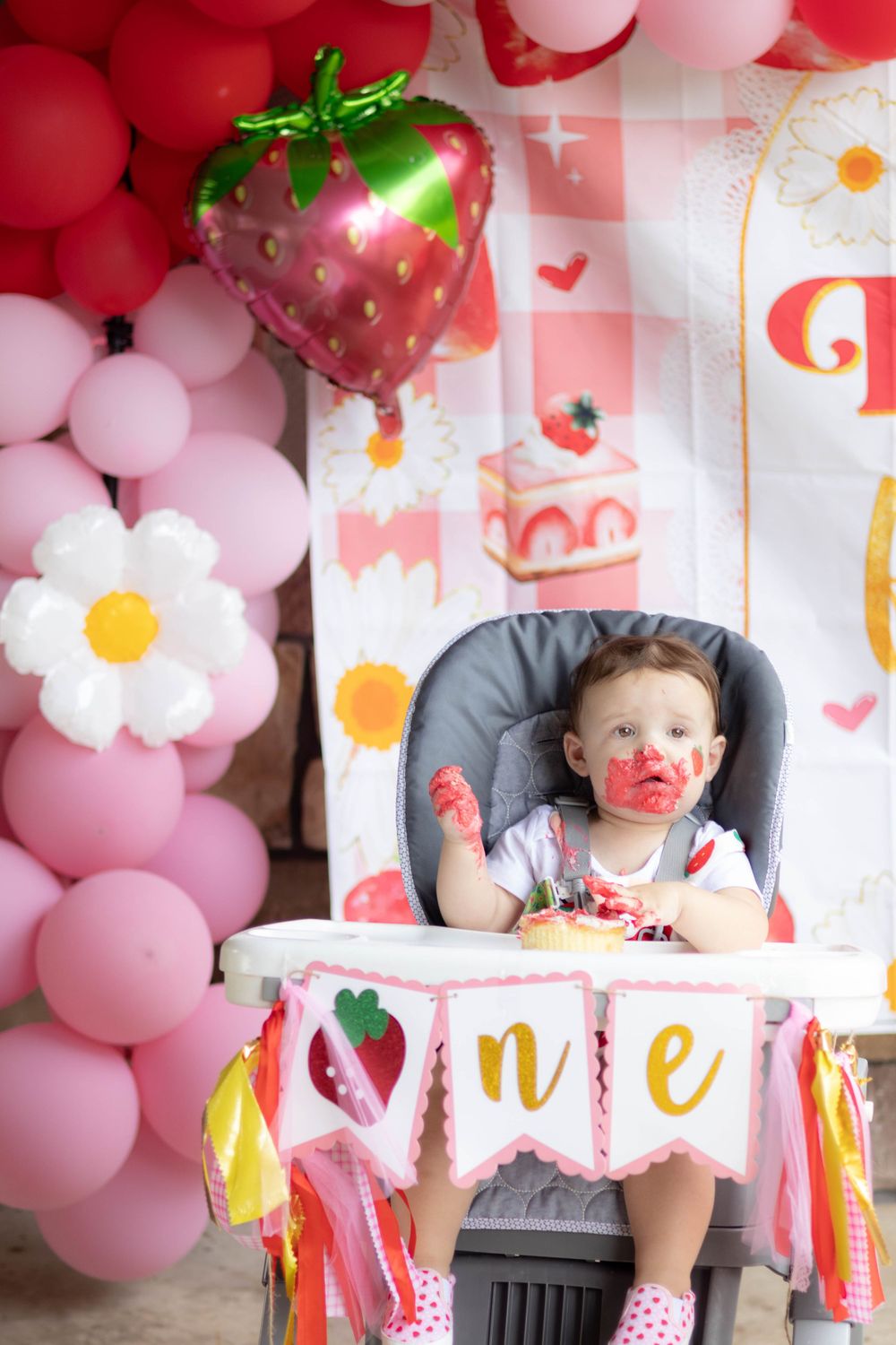 A first birthday party setup with pink balloons, strawberry decorations, and a high chair.