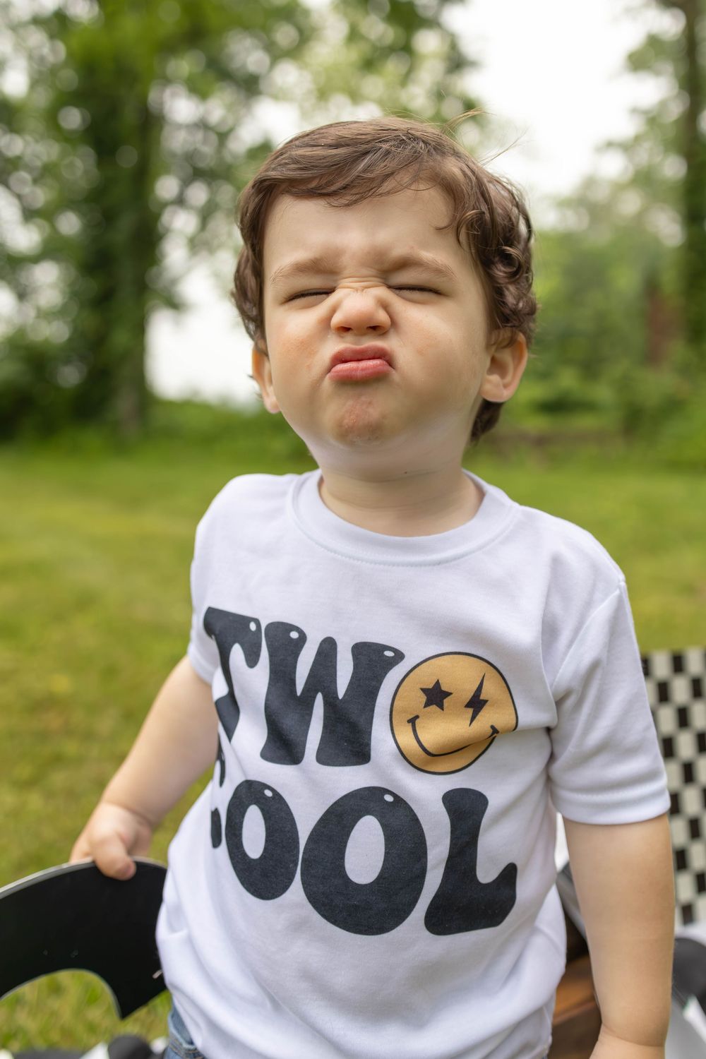 A toddler in a white graphic t-shirt with a smiley face design makes silly expressions in an outdoor setting.