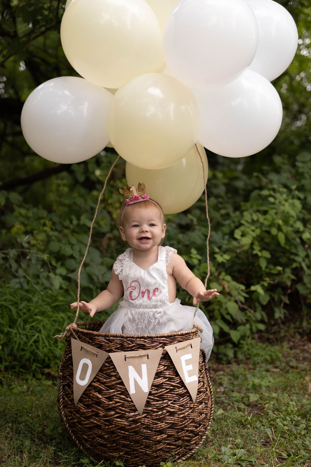 Baby in white dress sitting in woven basket with white balloons outdoors.