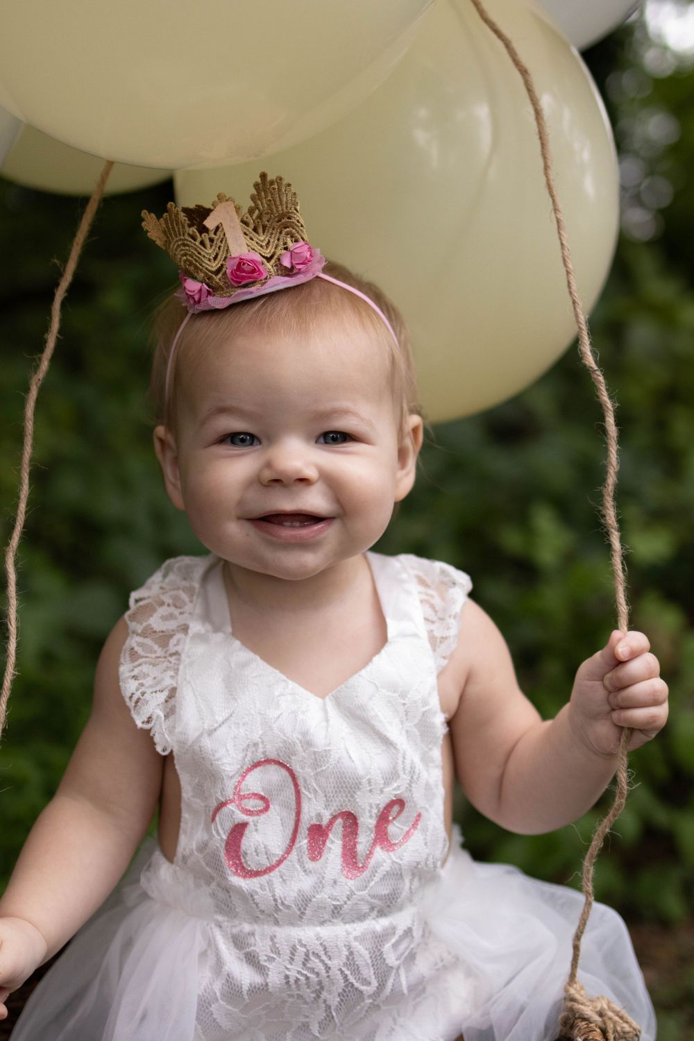 A happy child in a white dress and tiny crown holds white balloons during a celebration.