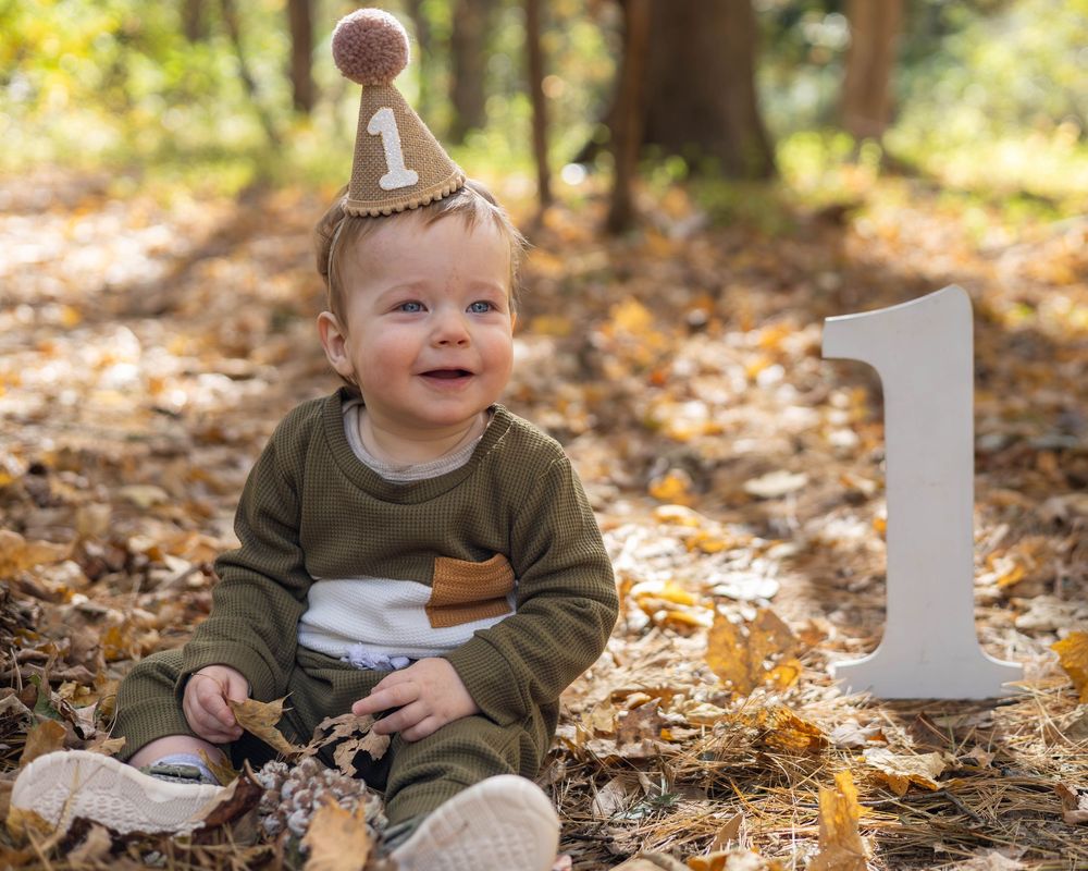A baby in a green sweater sits in autumn leaves during an outdoor photo session.