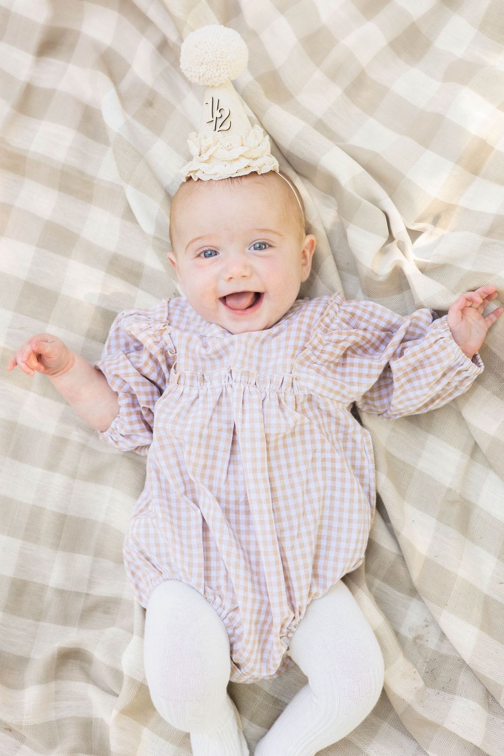 Baby in pink gingham dress lying on beige checkered blanket smiles joyfully while playing with white bow.