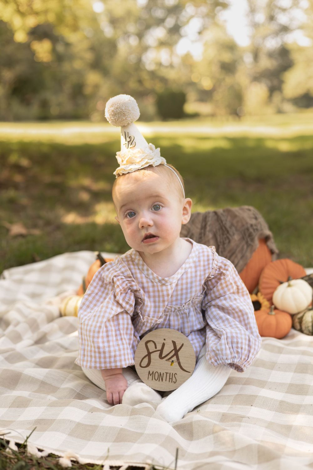 Baby wearing a birthday crown sits on plaid blanket with autumn pumpkins and decorations in outdoor setting.