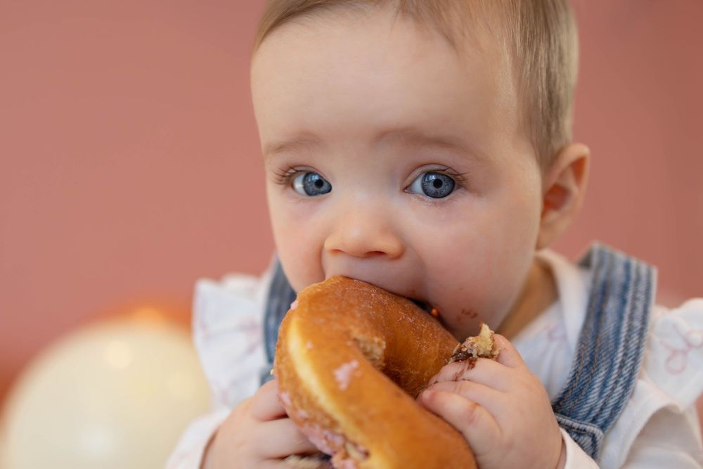Baby with bright blue eyes takes a big bite of a glazed donut against a pink background.