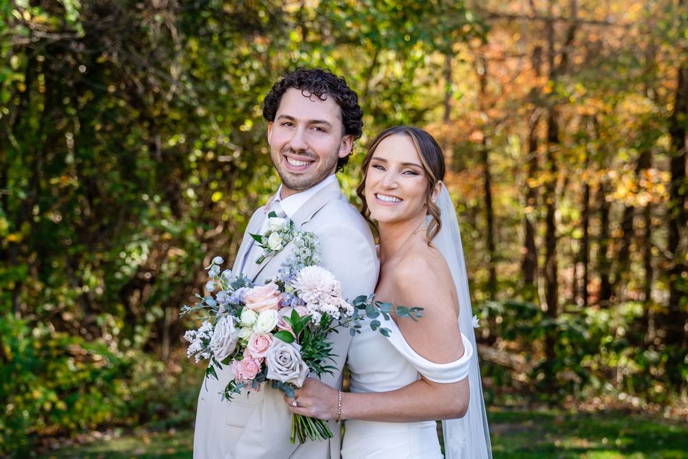 Smiling bride and groom pose together outdoors holding a pink and white bouquet with autumn foliage in the background.