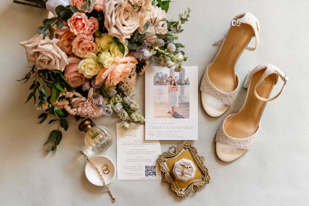 Elegant wedding flat lay featuring blush floral bouquet, lace heels, gold rings, perfume, and invitation suite on white surface.