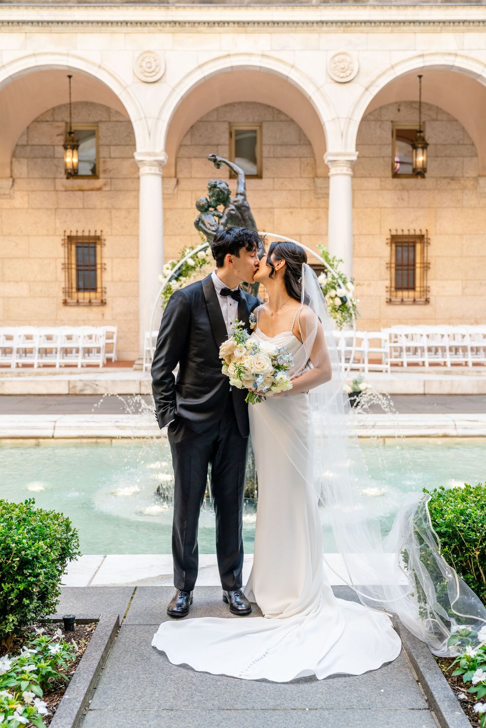 Bride and groom kissing by a fountain in an elegant courtyard with white chairs and floral arch at their wedding ceremony.