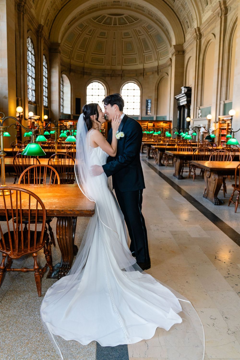 Bride and groom share a kiss in an elegant library with green lamps, wooden tables, and a grand vaulted ceiling.