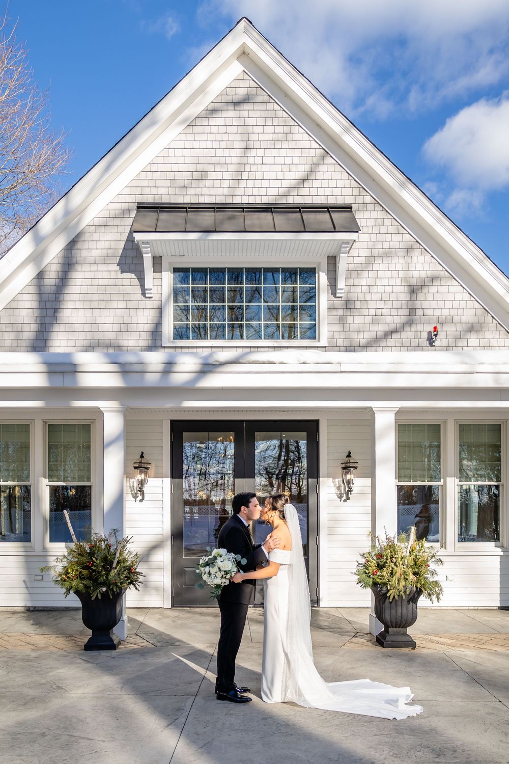 Bride and groom share a kiss outside a charming white shingle wedding venue under a bright blue sky.