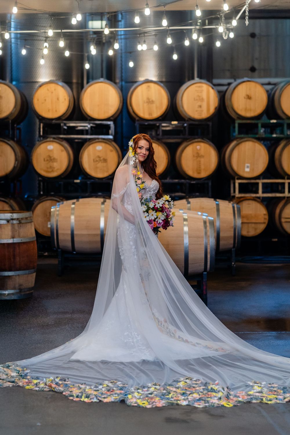 Bride in flowing white lace gown stands in front of rustic wine barrels in winery with hanging Edison bulbs at Labelle Winery Amherst NH