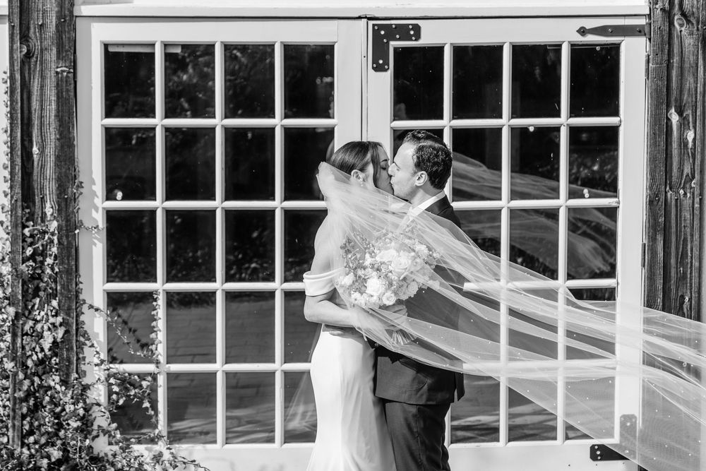 Black and white photo of a bride and groom sharing a kiss, with her veil flowing in the wind before a white French door.