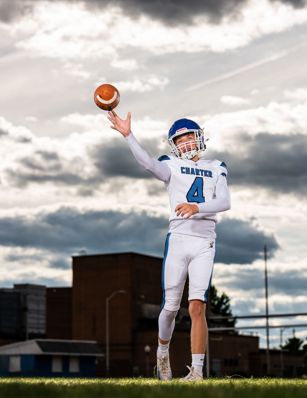 Football player in white uniform and blue helmet reaches up to catch ball against dramatic cloudy sky.