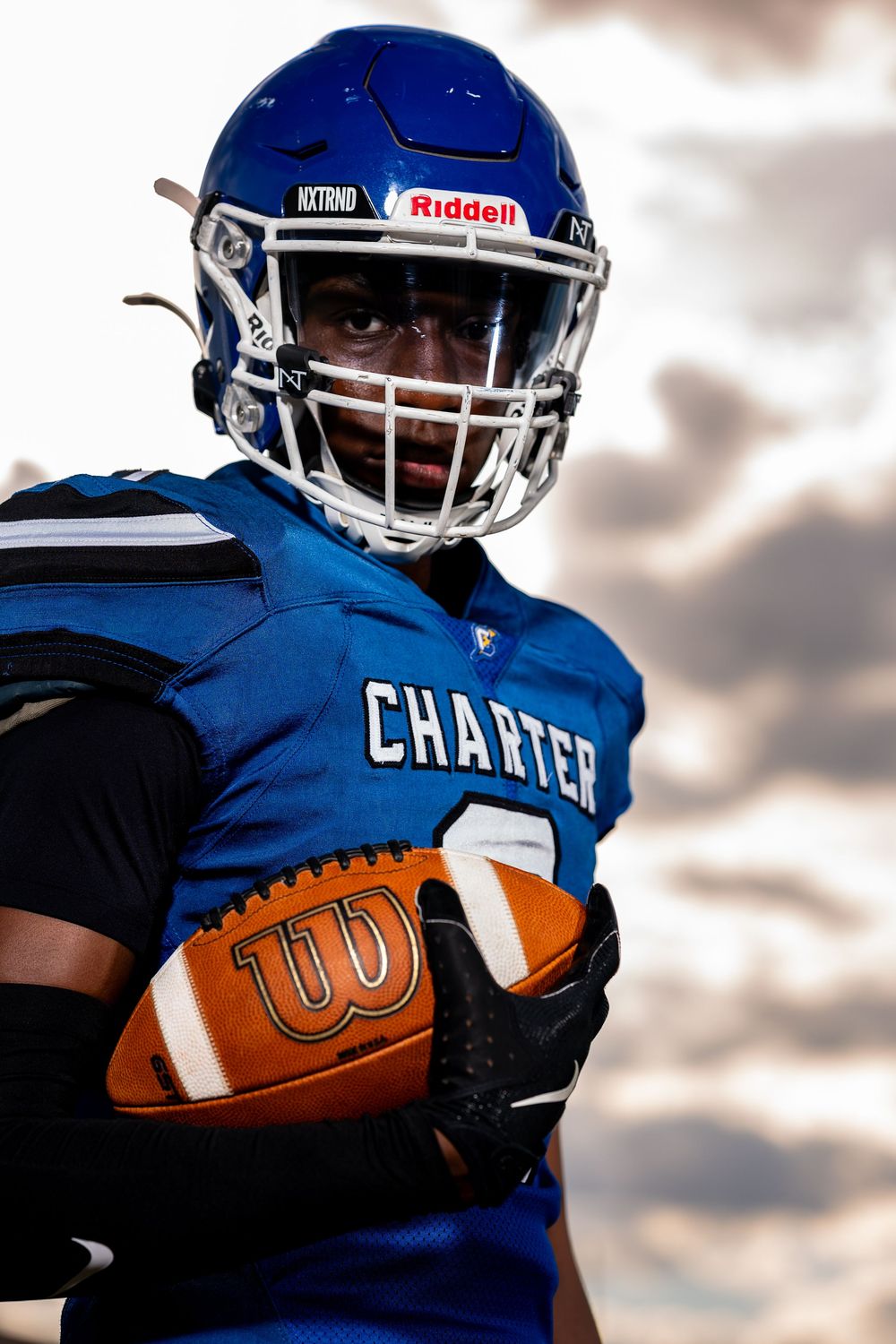 Football player in blue uniform carries ball under dramatic cloudy sky during game action.
