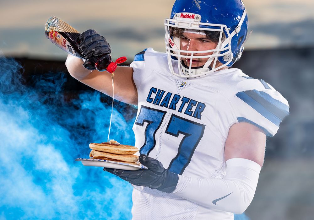 A football player in white and blue uniform poses dramatically with blue smoke in the background during a photoshoot.